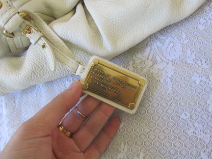 Hand holding a gold tag attached to a white leather bag on a textured white surface