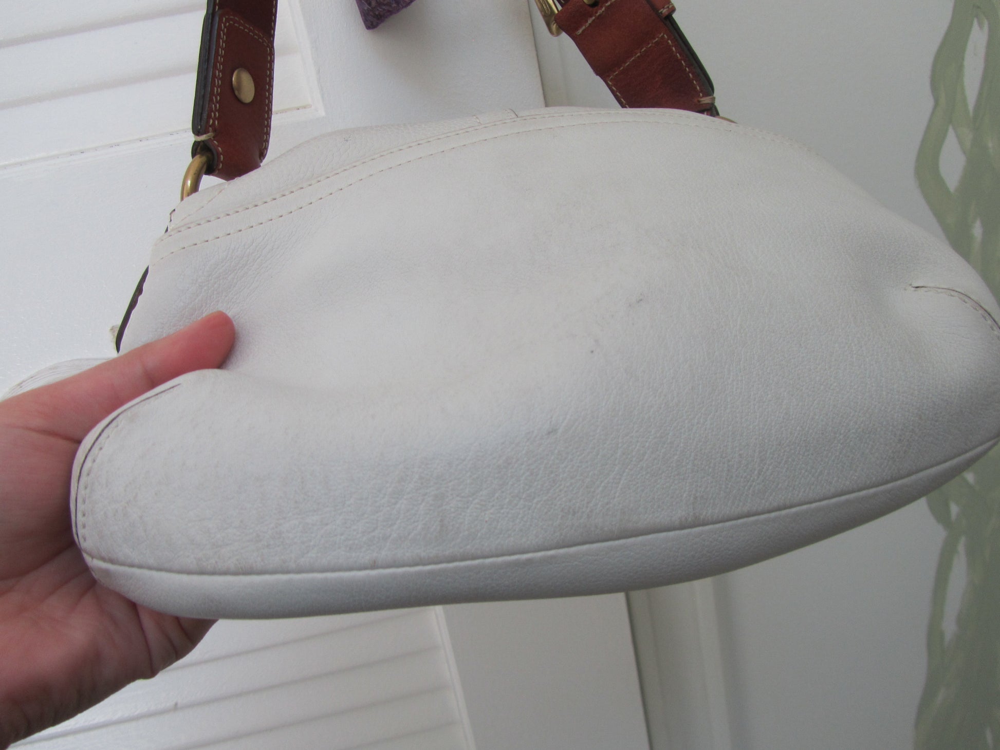 White leather handbag with brown strap held by a hand against a white background