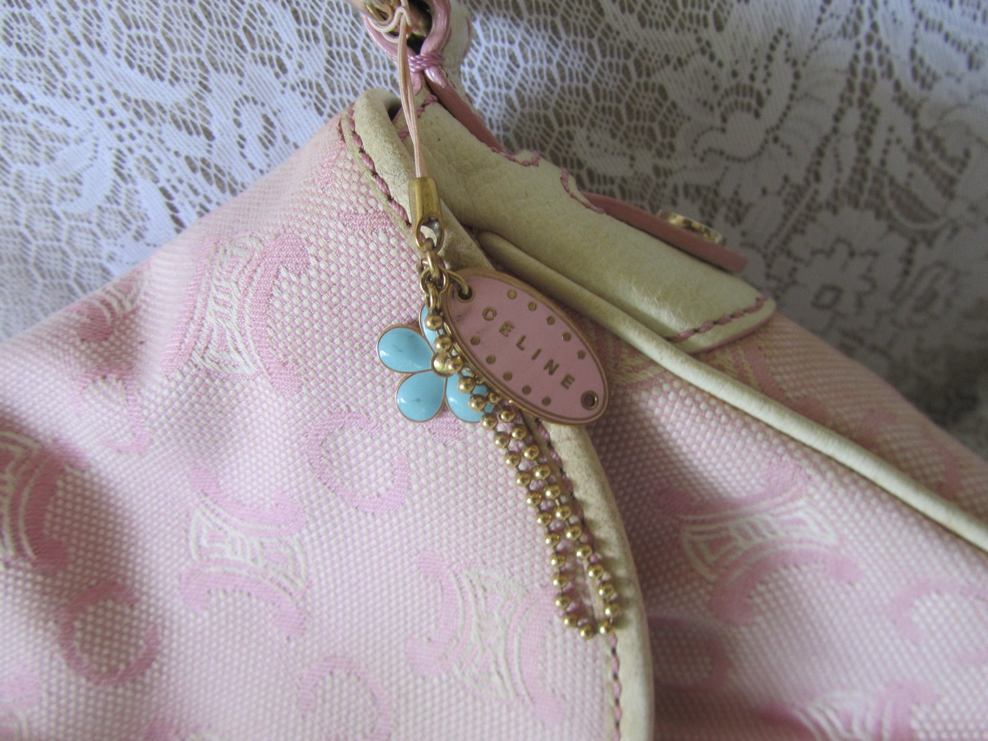 Close-up of a pink Celine handbag with a gold chain and blue flower charm on a lace background.